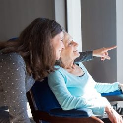 Positive mother and daughter enjoying dramatic view out of window. Young health visitor showing to senior lady funny scene out of window. Family relations concept